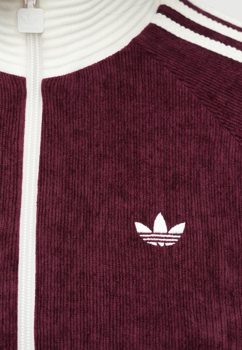 Bordeaux corduroy ritsjack met een witte ribgebreide kraag, voorzien van een klein wit logo aan de linkerkant en gestreepte accenten op de schouders.