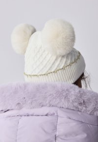 Fehér kötött sapka két nagy szőrme bojtokkal, aranyozott bordázott szegéllyel. Világos lila műszőrme galléros dzsekivel viselve.