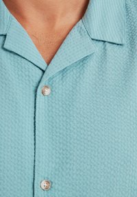 Light blue short-sleeve button-up shirt in textured fabric, featuring a pointed collar and pearl-like buttons on the front.