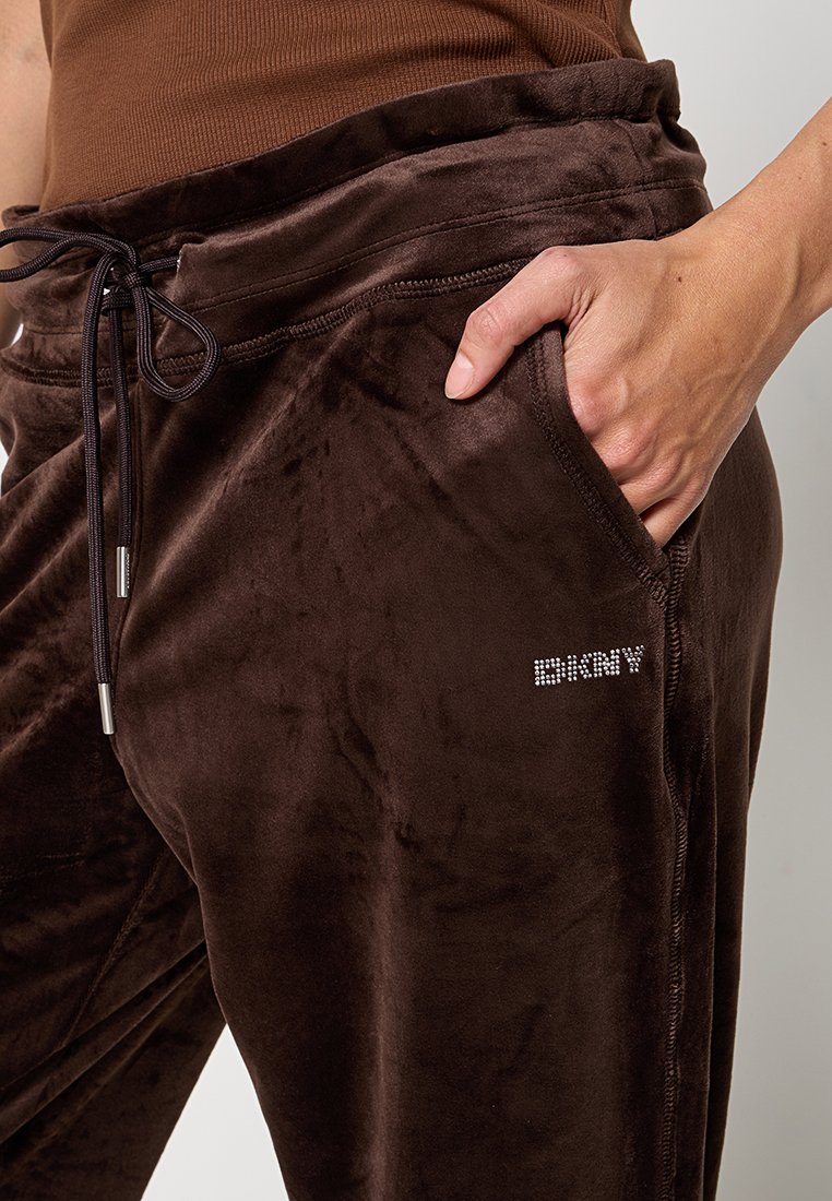 Aksamitne brązowe dresy z regulowanym paskiem w talii, bocznymi kieszeniami oraz akcentem z kryształków na lewej udzie. Gładka tekstura.