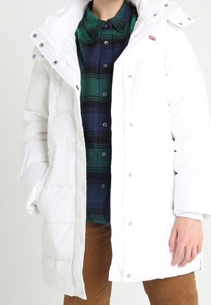 Person iført en hvid lang dunjakke med en grøn og marineblå ternet skjorte og brune bukser, hænder afslappet ved siderne og i lommerne.