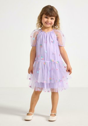 Giovane ragazza sorridente, che indossa un vestito viola chiaro con maniche a sbuffo e ricami floreali colorati, in piedi su uno sfondo bianco uniforme.