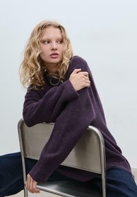 Lila, übergroßer Pullover aus weichem Material, mit weiten Ärmeln und geripptem Saum. Das Model sitzt auf einem silbernen Metallstuhl und trägt dunkle Jeans.