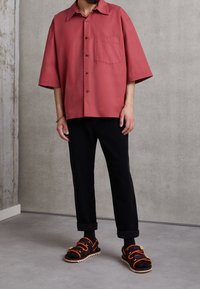 Brede shirt met korte mouwen in koraal met een borstzak, gecombineerd met zwarte corduroypantalons en zwarte sandalen met oranje banden en een zigzagzool.