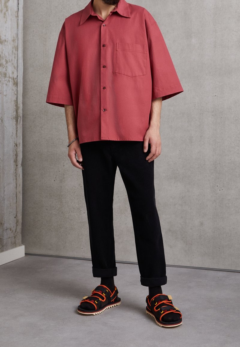 Brede shirt met korte mouwen in koraal met een borstzak, gecombineerd met zwarte corduroypantalons en zwarte sandalen met oranje banden en een zigzagzool.