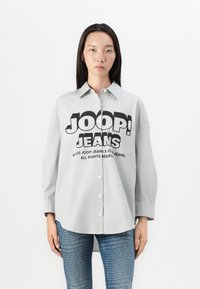 Stripet grå skjorte med lange ermer og en løs passform. Den har et stort svart "JOOP! JEANS" grafikk på forsiden. Kombinert med denimbukser.