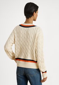 Kremasti džemper s pletenim kabelskim uzorkom i rebrastim rubom, s narančastim i mornarsko plavim prugama na izrezu i donjem dijelu. Dugih rukava, ležernog kroja.