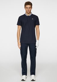 Marinblå pikétröja med stickad krage och treknappsslutning, matchad med byxor av cargo-modell och vita sportskor, bärs av en stående manlig modell.