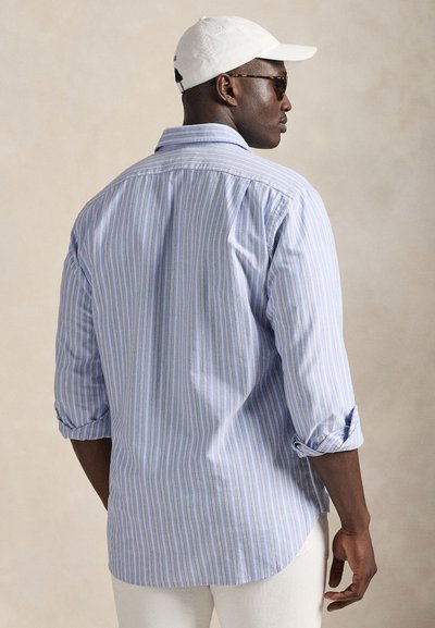 Blå och vit randig skjorta med knappar fram och uppkavlade ärmar, lätt tyg, ryggparti med yoke-detalj, matchas med en vit keps och solglasögon.