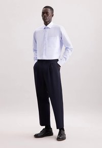 Lichtblauw, langärmelig overhemd met knopen en een borstzak, gecombineerd met donkere geplooide broek en zwarte schoenen, staande tegen een neutrale achtergrond.