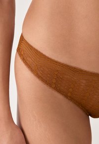 Nahaufnahme der Hüfte einer Person, die rustfarbene Spitzenunterwäsche mit geometrischen Mustern auf glatter Haut trägt.