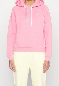 Rosa huvtröja gjord av bomullsblandning, med en känguruficka, vita dragsnören och ribbade muddar och fåll. Minimal logotypdetalj.