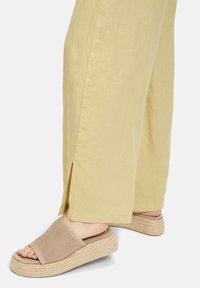 Beige schuifsandalen met een zachte suede bovenkant, een dikke geweven espadrille zool en een iets verhoging platform, gecombineerd met lichtgele linnen broek met zijsplitten.