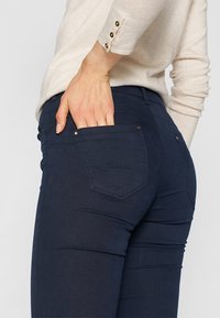 Pantalones de mezclilla azul oscuro con un diseño ajustado. Las características notables incluyen bolsillos traseros y herrajes en tono dorado. Textura suave, silueta ajustada.
