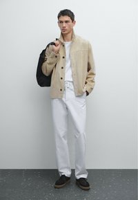 Beige suede jas met drukknoopjes, witte T-shirt en witte broek. Zwart rugzak, donkere schoenen met rubberen zolen. Effen gestructureerde achtergrond.