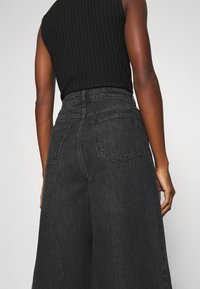 Musta, korkeavyötäröiset leveälahkeiset denimpantsarit, joissa on teksturoitu kangas, näkyvät takataskut ja joustava vyötärönauha. Yläosana musta ribbikuvioinen hihaton toppi.