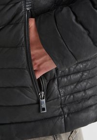 Schwarze Lederjacke mit gesteppter Textur, sichtbarem Reißverschlussfach, schlankem Design und glatter Oberfläche. Mit silbernen Beschlägen.