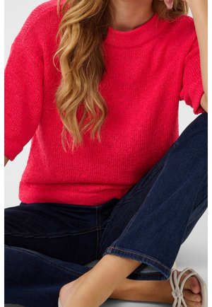 Femme aux longs cheveux ondulés portant un pull en maille rouge vif, un jean bleu foncé et des sandales blanches à bout ouvert, assise avec une jambe croisée.