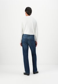 Hombre vestido con una camisa blanca de manga larga, jeans azules y zapatos negros se encuentra de pie frente a una pared blanca con las manos a los lados.