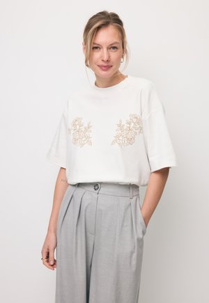 Jonge vrouw met blond haar draagt een wit T-shirt met beige bloemenprints en hoge taille lichtgrijze geplooide broek, staand met hand in de zak.