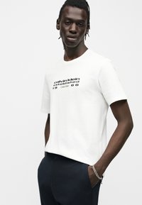Νέος άνδρας που φοράει λευκό μπλουζάκι Calvin Klein με μαύρο κείμενο "calvin klein established 1968" και σκούρο παντελόνι, χέρια στις τσέπες, ουδέτερο φόντο.