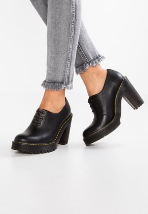 Personne portant des chaussures en cuir noires épaisses à talons hauts lacées avec des coutures jaunes et un jean gris à ourlet effiloché sur un sol blanc.
