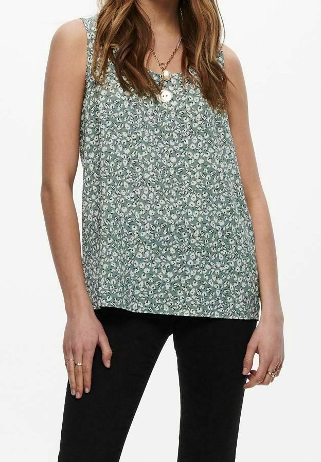 Femme portant un haut sans manches vert clair à motif floral avec deux colliers en or et un pantalon noir, debout dans une posture détendue.