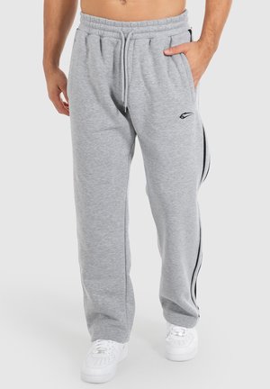 Man wearing light gray sweatpants with black side stripes and white sneakers, standing with one hand in pocket against white background.