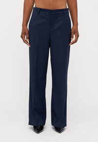 Calças largas azuis-marinho feitas de um tecido suave, com frente lisa, passantes para cinto e ajuste sob medida. Sapatos pretos de bico fino visíveis.