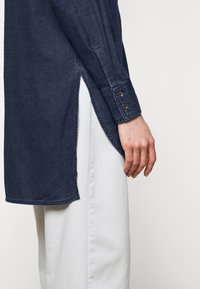 Camicia di jeans blu navy con maniche lunghe, fessure laterali e polsini con bottoni, abbinata a pantaloni bianchi a gamba larga. Texture liscia, vestibilità rilassata.