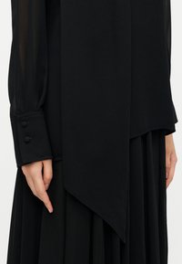 Μαύρη μπλούζα με μακριά διάφανα μανίκια και ασύμμετρο τελείωμα. Διαθέτει μανσέτες με κουμπιά και λείο ύφασμα με ρέουσα σχεδίαση.