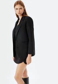Schwarzer maßgeschneiderter Blazer mit zwei Knöpfen, strukturierten Schultern und Fronttaschen, kombiniert mit einem schwarzen Mini-Kleid und knielangen Stiefeln.
