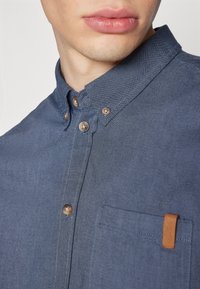 Blauwe, knoopsluiting overhemd met een kraag, textuurstof en een borstzak met een bruine leren accenten nabij de stiksels.