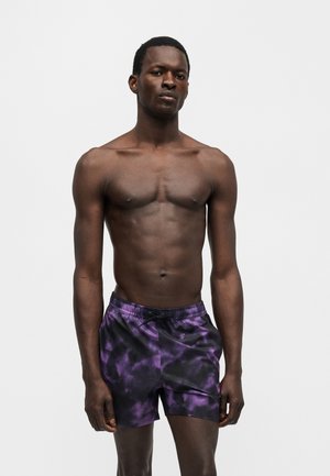 Homme torse nu à la peau foncée portant un short de bain tie-dye violet et noir sur un fond gris clair uni.
