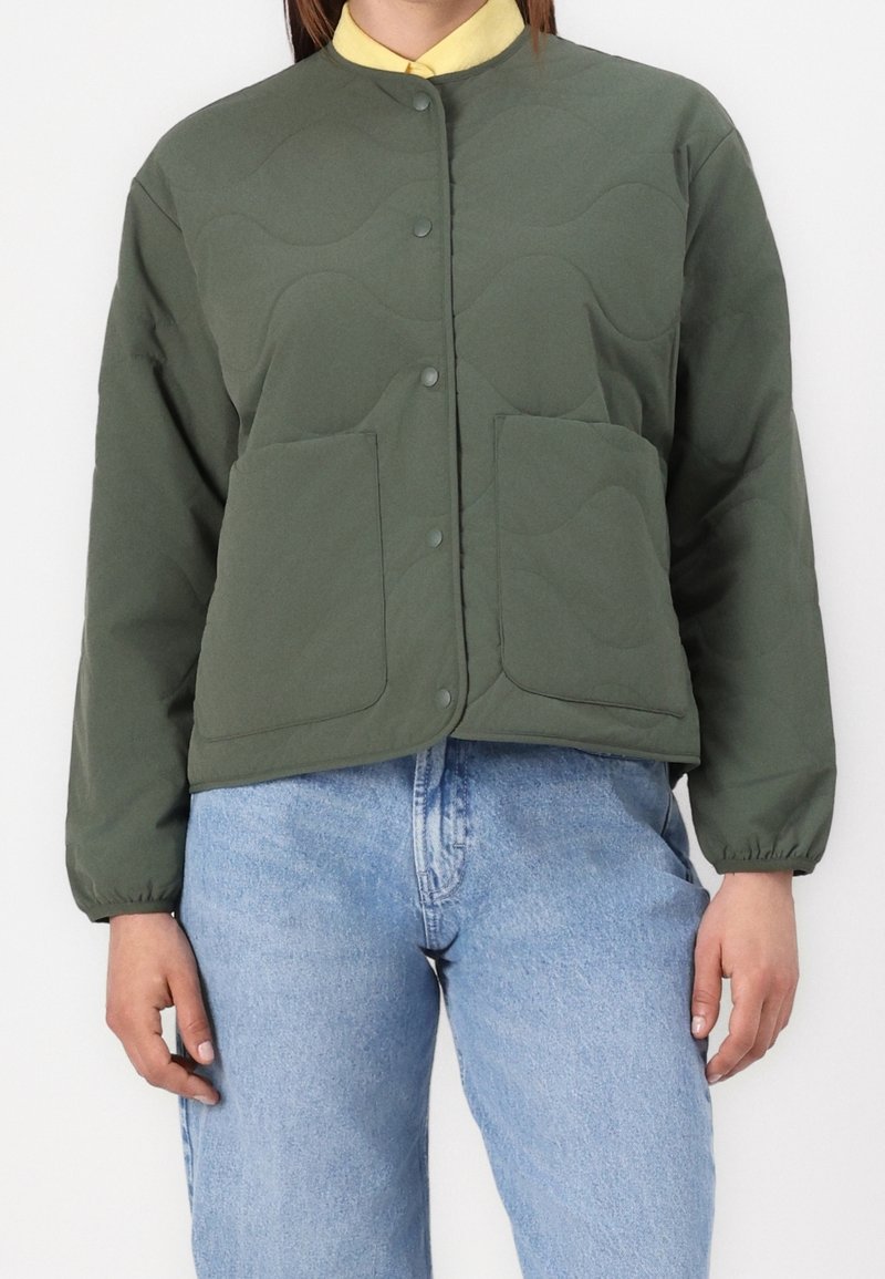 Personne portant une veste matelassée verte avec de grandes poches avant, sur une chemise jaune à col et un jean bleu clair, debout devant un fond blanc.