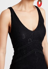 Černé krajkové šaty s hlubokým výstřihem do V, mají pevný střih a křížící se texturované pruhy. Zahrnují výrazné červené kruhové náušnice.