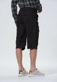 Sorte cargo shorts lavet af slidstærkt stof, knælange med sidelommer og en afslappet pasform. Kropsbåret med lysegrå sneakers med brune accenter.