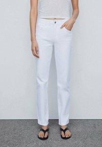 Jeans de mezclilla blanca con un corte recto, diseño de cinco bolsillos y cierre de botón. Combinados con sandalias negras y una blusa ajustada.