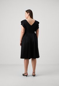 Vestido envoltorio negro con mangas cortas con volantes, espalda en V y cuerpo ajustado. La falda acampanada cae hasta la mitad de la pantorrilla, combinada con simples tacones negros.