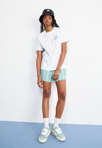 White t-shirt with a blue circular logo, light green and blue striped shorts, and mint green sneakers with white laces. Black bucket hat.