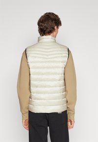 Beige Weste mit Padding, hohem Kragen und horizontal gestepptem Design, kombiniert mit einem tanfarbenen Langarmshirt und schwarzen Hosen.