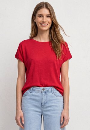 Giovane donna sorridente, con indosso una maglietta rossa a maniche corte e jeans azzurri, in piedi davanti a uno sfondo grigio chiaro uniforme.