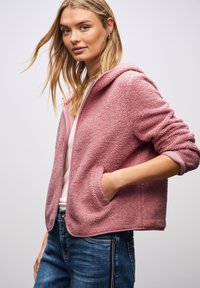 Młoda kobieta z długimi blond włosami, ubrana w teksturowany różowy polar na zamek z kieszeniami, białą koszulę i niebieskie dżinsy, na jednolitym tle.