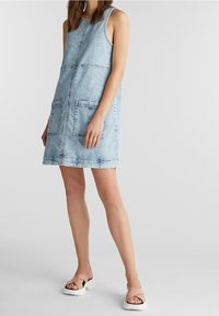 Vaaleansininen farkkumekko, jossa on kaksi etutaskua, olkaimeton muotoilu ja näkyvät ompeluyksityiskohdat. Yhdistetty vaaleanbeigeihin hihnakenkiin.