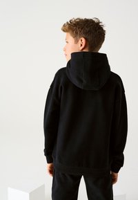 Must kapuutsiga dressipluus lõdva lõikega, millel on soonikkoes varrukad ja alläär. Pehme tekstuur, lihtne disain ja silmapaistev kapuutsi detail.