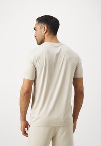 Camiseta ajustada beige de manga corta, con textura suave, cuello redondo y detalles de costura, vista por la espalda de una figura masculina.