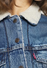 Blå jeansjacka med vit sherpakrage. Har metallknappar och kontrastsömmar. Märkbart rött logomärke på sidofickan.