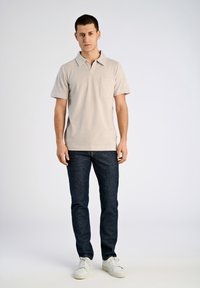 Beżowa koszulka polo z krótkim rękawem i kieszonką na piersi, zestawiona z ciemnoniebieskimi dżinsami slim fit i białymi sneakersami, stojąca na jednolitym tle.