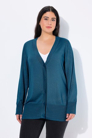 Frau trägt eine türkisfarbene, geknöpfte Strickjacke über einem weißen Top und schwarzen Hosen, steht vor einem schlichten weißen Hintergrund.