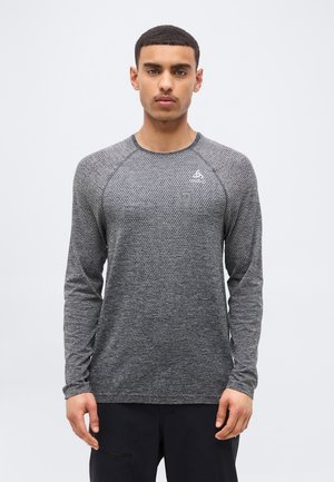 Νεαρός με κοντά μαλλιά, που φοράει γκρι αθλητική μπλούζα με υφή και μαύρο παντελόνι, στέκεται μπροστά σε απλό φόντο.